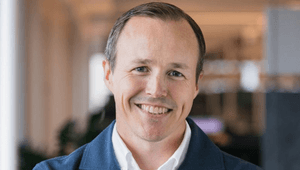 Ett närporträtt av en leende man med kort ljusbrunt hår. Han bär en vit skjorta under en blå, texturerad kavaj eller kofta. Bakgrunden är ljus och mjukt suddig, vilket ger bilden en professionell känsla.