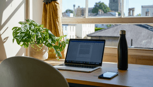 Inomhusbild som visar ett brunt träbord framför ett fönster. En silverfärgad bärbar dator med ett öppet dokument och en svart termosflaska står på bordet. Till vänster står en grön krukväxt. Utanför fönstret syns hustak och ett gammalt kyrktorn i bakgrund