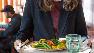 Närbild av en person som bär en bricka med en måltid bestående av ris, grönsaker samt ett glas vatten i en restaurangmiljö.