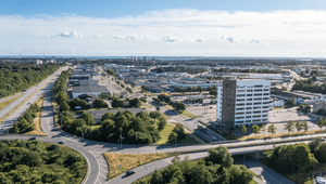 Högt flygfoto över ett industri- och handelsområde med en modern, ljus kontorsfastighet, trafikerade vägar och grönområden under en klarblå sky
