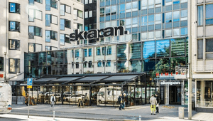 Exteriörbild i ögonhöjd över entrén till gallerian och skyskrapan Skrapan med inglasad uteservering, fasadskyltar och fotgängare.