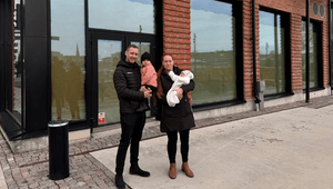 En man och en kvinna står utomhus framför en modern tegelbyggnad med stora glaspartier. Båda håller i varsitt barn, varav ett är en baby i en vit overall. De är klädda i mörka vinterjackor och miljön ser ut som ett nybyggt område med stenlagd mark.