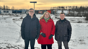 Tre personer står uppställda framför ett snötäckt fält i skymningen, med en kvinna i röd kappa i mitten flankerad av två män i mörka vinterjackor.