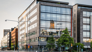 Exteriör av en modern kontorsbyggnad i glas och mörk sten vid Hjälmarekajen. I den stora glasfasaden reflekteras ett äldre torn, och framför byggnaden syns gröna träd, gatubelysning och en tom väg i mjukt dagsljus.