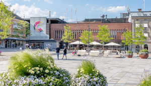 Ett öppet stadstorg med ljus stenläggning framför köpcentret "Commerce". Byggnaden har en tegelfasad och en stor färgglad logotyp. På torget finns moderna sittmöbler, stora blomsterarrangemang i lila och gult, samt träd. Människor promenerar och sitter vi