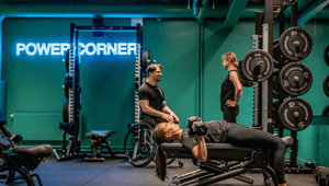 En kvinna gör hantelpress på en träningsbänk i ett gym med turkosa väggar och neonskylten "POWER CORNER".