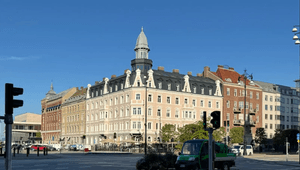 Exteriörbild i grodperspektiv över ett stadslandskap med en klassisk hörnbystnad med torn, omgiven av trafik och korsning under en klarblå sky.