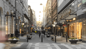 Fotgängare promenerar längs en stensatt gågata kantad av butiker, fasadbelysning och blomsterurnor i mjukt dagsljus.