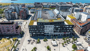 Flygfoto över ett torg och stadsområde med en stor mörk fastighet utrustad med solpaneler och växttak vid havet.