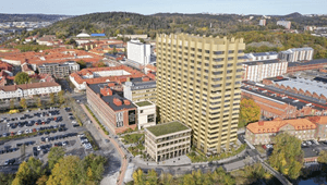 Här är en beskrivning av bilden i löpande text:  En flygfoto-rendering visar ett stort, högt byggnadskomplex i en tät stadsmiljö, omgivet av äldre byggnader och grönskande kullar. I centrum står ett höghus i ljust gulbeige färg med en distinkt, räfflad fa