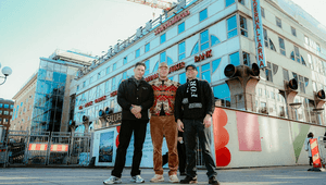 Tre män står uppställda framför Medborgarplatsen och Söderhallarna i Stockholm. Byggnaden är delvis täckt av byggställningar och plank, vilket signalerar pågående renovering, under en klarblå himmel.