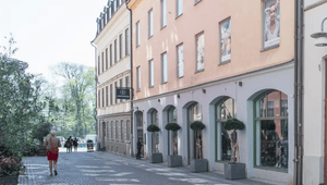 En kullerstensgata i en stadsmiljö flankerad av byggnader i pastellfärger, med en klinik för laser och estetik i förgrunden och fotgängare som promenerar i solljuset.