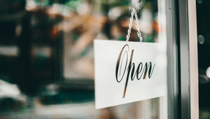 En närbild av en vit skylt med texten "Open" i kursiv stil, som hänger i ett fönster eller en glasdörr. Skylten hänger i en metallkedja som fästs med en sugpropp. Bakgrunden är suddig och visar gröna och bruna nyanser,