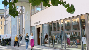 Kontorsentré på Kils gatan 4 med glasfasad. Människor passerar utanför i solsken medan trädgrenar med gröna blad ramar in bilden.