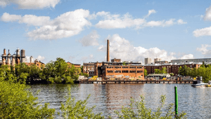 En vy över ett vattennära industriområde med äldre tegelbyggnader och en hög skorsten under en blå himmel med vita moln. I förgrunden syns glittrande vatten och gröna träd som ramar in bilden.