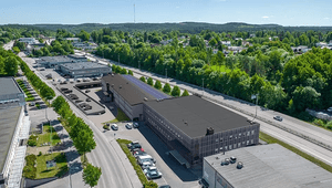 Flygfoto över ett industri- och kommersiellt område med långsträckta fastigheter, parkeringsplatser och en intilliggande bilväg omgiven av lummig grönska.