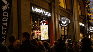 Kvällsbild på en Bastard Burgers-restaurang med upplysta skyltar och en folksamling som väntar utanför entrén.