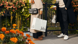 En närbild på två personer som står vid ett broräcke dekorerat med rikliga blomsterarrangemang i orange och rosa. Den ena personen bär en beige kappa, en svart axelremsväska och håller i en vit shoppingkasse med texten "Johnells". Den andra personen bär e
