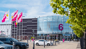 Väla köpcentrums entré nummer 6 med en stor rundad glasfasad, flaggor som vajar i vinden och parkerade bilar i förgrunden.