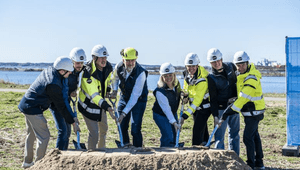 En grupp människor i byggvästar och skyddshjälmar tar ett gemensamt första spadtag för ett byggprojekt. De står utomhus vid en sandhög med havet i bakgrunden under en klarblå himmel.