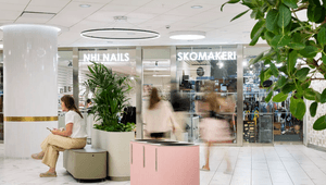 Ljus galleriagång med butikerna NHI Nails och Skomakeri, en sittande kvinna samt en rosa källsorteringsmöbel.