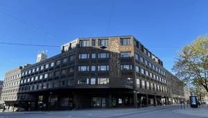 En mörk, flervånings kontorsbyggnad i sten med en utskjutande arkad i gatuplan, belägen i en vägkorsning under en klarblå himmel. Byggnadens fasad pryds av ett vertikalt konstverk i relief, och enstaka fotgängare syns på trottoaren vid byggnadens bas.