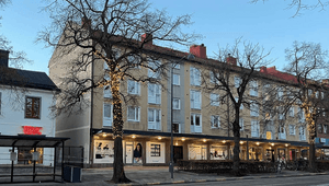Bild tagen under kvällstid av ett bostadshus med tre våningar i ljusgul puts. På gatuplanet finns flera butiker under ett utskjutande tak med inbyggd belysning. Flera kala träd står framför huset, varav ett är dekorerat med julbelysning