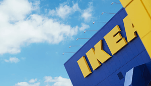 Närbild i grodperspektiv på en blå IKEA-varuhusfasad med den gula logotypen mot en blå sky med vita moln.