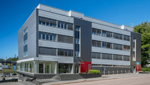 Modern kontorsbyggnad i fyra våningar med grå fasadpaneler, röda accentdetaljer och entrétrappa mot gatan under en klarblå sky.