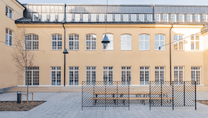 En stenlagd innergård framför en klassisk, ljusgul byggnad med regelbundna fönsterrader, bänkar och svarta metallskärmar.