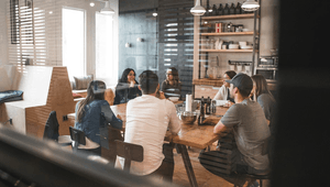 En grupp människor sitter samlade runt ett långt träbord i en avslappnad kontorsmiljö eller fikarum. De samtalar i ett ljust rum med industriell känsla, där en öppen hylla med porslin och stora fönster syns i bakgrunden.