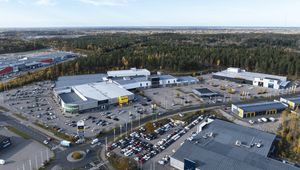 Flygfoto över ett handelsområde med stora butikskomplex, parkeringsplatser och en rondell. Bland butikerna syns skyltar för Rusta och Second Hand, omgivna av vidsträckt skog och mark under en blå himmel med lätta moln.