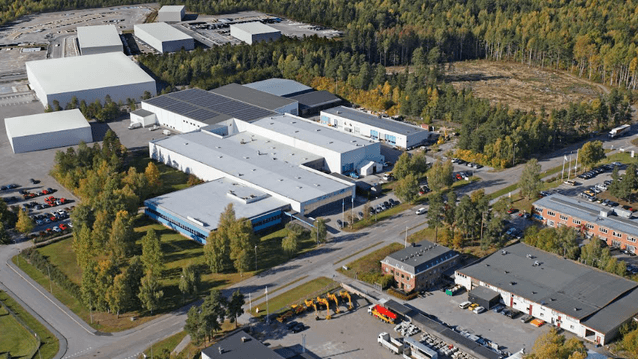 Flygfoto över ett industriområde med stora lagerbyggnader och verkstäder omgivna av skog och parkeringsplatser i höstfärger.