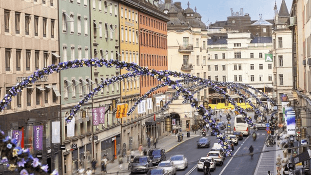 En livlig stadsgata med färgglada husfasader i grönt, gult, orange och beige. Tvärs över gatan hänger bågar dekorerade med blå och lila blommor. Bilar, cyklar och människor rör sig längs gatan, medan skyltar och butiker kantar trottoarerna.