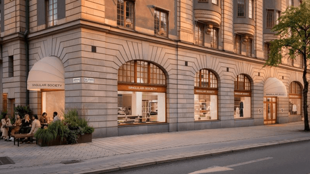 En ståtlig stenbyggnad i klassisk arkitektur med butiken "Singular Society" i gatuplan. Fasaden har stora välvda fönster och ljusa markiser. Till vänster finns en liten uteservering med människor som sitter vid ett bord omgivna av gröna växter. Gatan utan