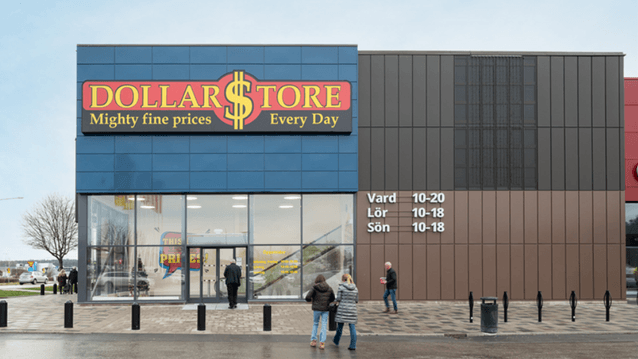 Exteriör av en DollarStore-butik med stora glaspartier vid entrén, tydliga öppettider på fasaden och människor som rör sig utanför.