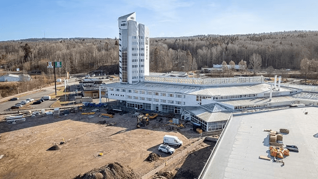 Vy över Navet i Jönköping under pågående markarbete och ombyggnation. Den vita byggnaden med sitt ikoniska torn syns bredvid en stor byggarbetsplats, med skogsklädda höjder i bakgrunden.