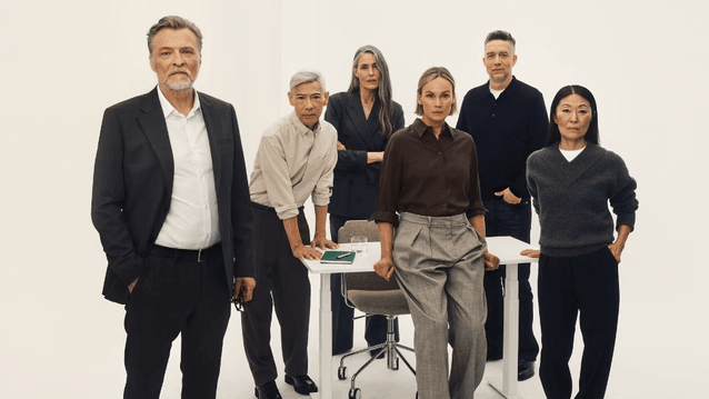 Ett gruppfoto av sex personer i affärskläder som står samlade kring ett vitt skrivbord mot en minimalistisk vit bakgrund. Gruppen har ett allvarligt och professionellt uttryck och bär kläder i sobra färger som svart, grått, brunt och vitt.