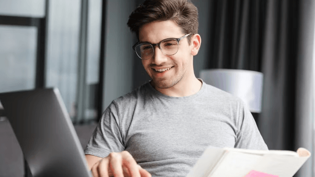 En leende ung man med glasögon som sitter vid en bärbar dator och studerar eller arbetar med en bok framför sig.