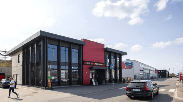 Exteriörbild i ögonhöjd över en modern snabbmatrestaurang med svart och röd fasad, stående bilar och en gående person.