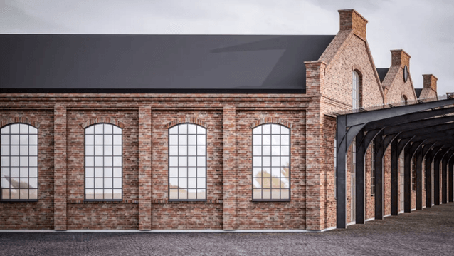 En lång industribyggnad i rött tegel med karaktäristiska bågfönster. Byggnaden har ett mörkt tak och en modern utbyggnad i form av ett svart stålskelett som skapar en övertäckt gångväg längs med fasaden. Markytan består av mörk gatsten.