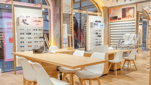 Interiör från en ljus och modern optikbutik med ett långt bord i ljust trä och vita stolar i förgrunden. Längs väggarna finns välexponerade hyllor med rader av glasögonbågar och prisskyltar som anger 'från 999 SEK'. Stora valvformade fönster i bakgrunden 