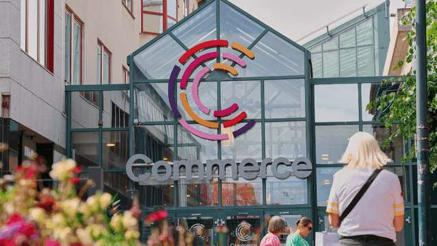 Entrén till köpcentrumet Commerce med en färgstark C-logotyp på glastaket och besökare i förgrunden.