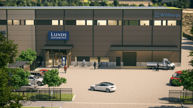 En modern brun byggnad med skyltar för Lunds universitet och Wihlborgs på fasaden. Utanför entrén syns människor som går eller cyklar, samt flera parkerade bilar och en liten lastbil. Byggnaden har stora fönster på andra våningen och omges av ett staket o