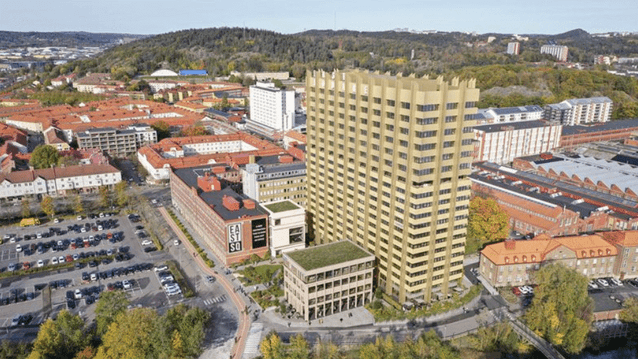 Flygfoto över ett stadsområde med en framträdande guldfärgad höghusbyggnad vid ett vattendrag. Omgivningen består av både äldre tegelbyggnader och grönska.