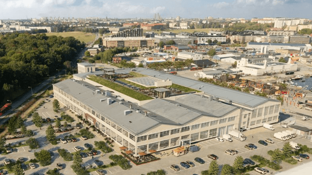 Flygfoto över en stor modern kontorsfastighet i ett industriområde, med en grönskande takterrass och stadssiluett i bakgrunden