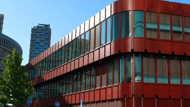 Modern byggnad i grodperspektiv med rödbrun plåtfasad, rundade hörn och stora fönsterband under en klarblå himmel.