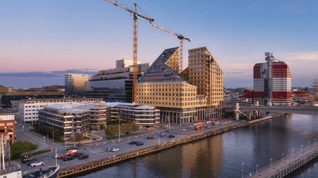 Flygfoto över ett hamnområde under byggnation med en spektakulär, trappstegsformad träbyggnad och en stor tornkran i skymningsljus.
