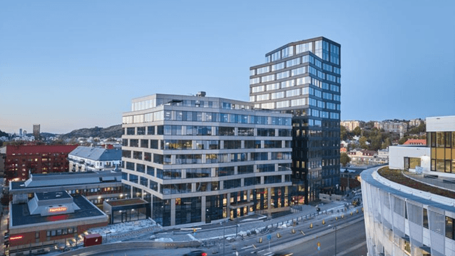 Ett modernt kontorskomplex med två stora glasbyggnader, varav en högre tornliknande byggnad, står intill en trafikerad rondell. Byggnaderna har många fönster och speglar himlen i glasfasaderna. Framför syns bilar som kör runt rondellen, och i bakgrunden l