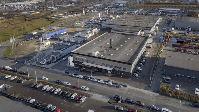 Flygfoto över ett modernt industri- eller handelsområde. I mitten syns en stor grå byggnad med platt tak, och texten "STRIDSYXAN 7" över huvudingången. Byggnaden är omgiven av stora parkeringsplatser fyllda med bilar, samt andra kommersiella fastigheter o